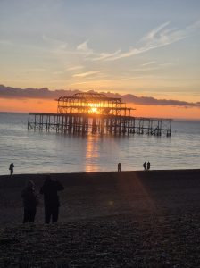 Sunset at Brighton’s West Pier on New Year’s Eve, with the skeletal remains of the pier silhouetted against an orange and gold sky, the sun reflecting on the calm sea, and small groups of people on the pebble beach watching and photographing the scene.