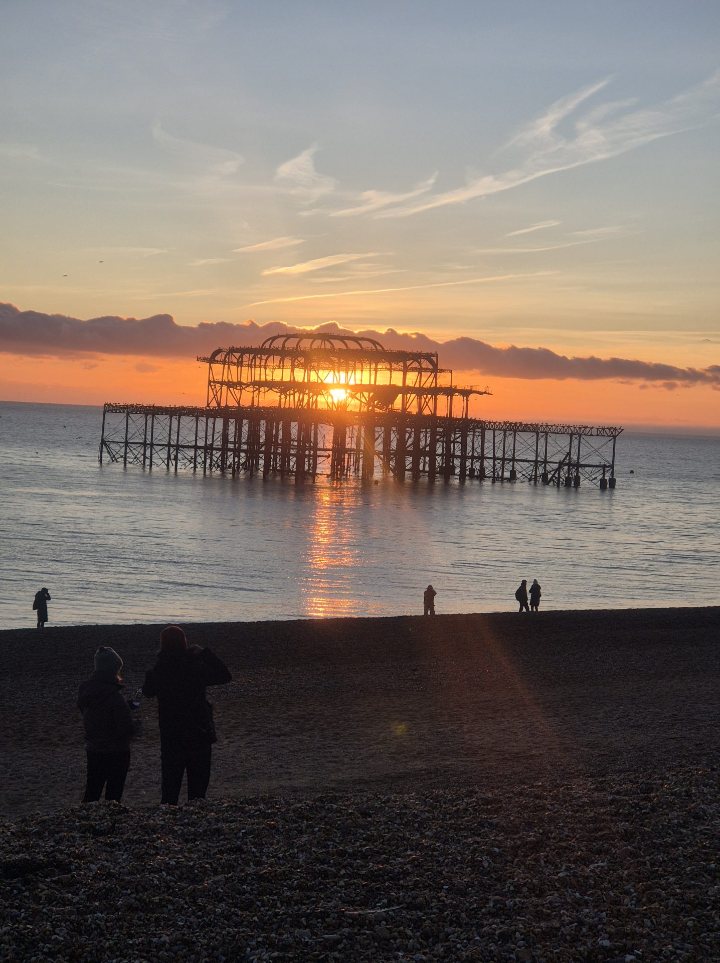Sunset at Brighton’s West Pier on New Year’s Eve, with the skeletal remains of the pier silhouetted against an orange and gold sky, the sun reflecting on the calm sea, and small groups of people on the pebble beach watching and photographing the scene.